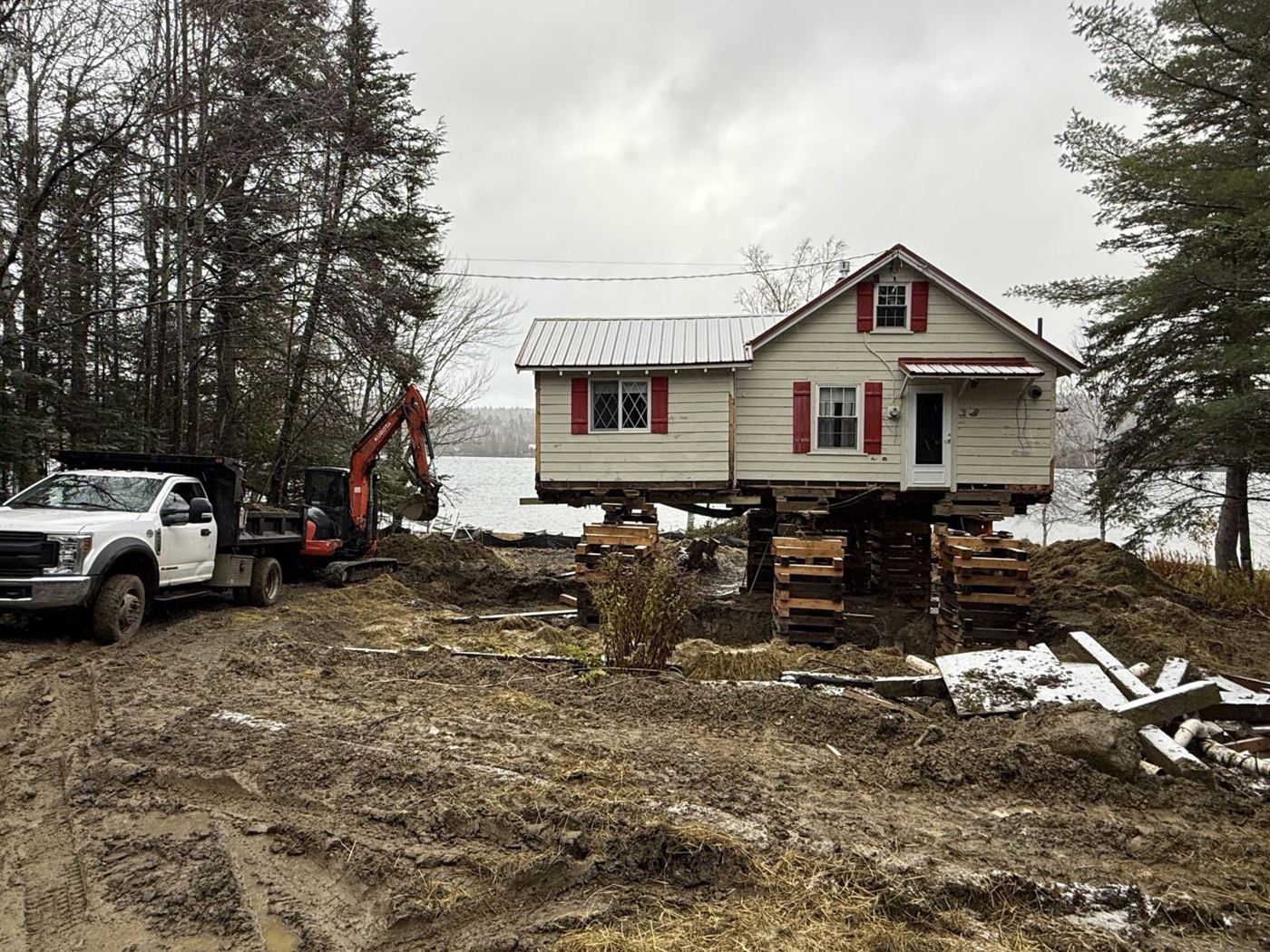 Red-shuttered lakeside camp lifted on cribbing, with truck and excavator on site