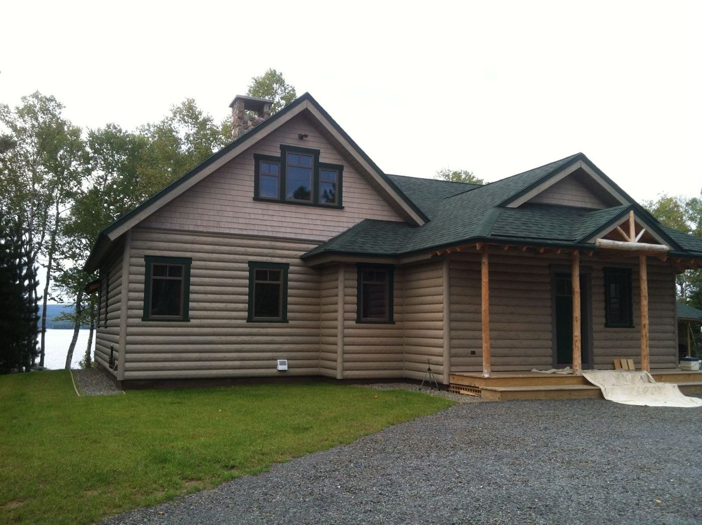 The finished home with log siding, green trim, stone chimney and a log-post porch, with the lake behind