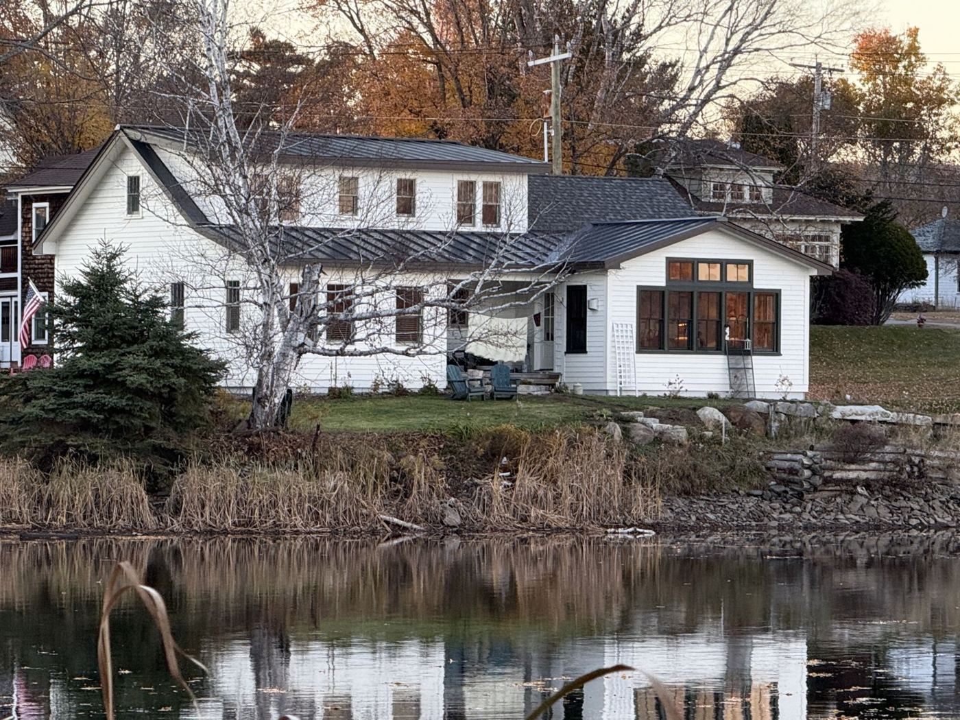 White lake house with a metal roof, seen across the water in fall
