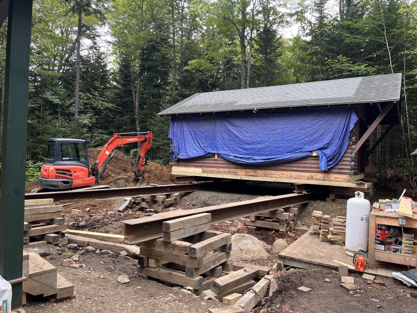 Log building raised on steel beams and cribbing, with an excavator on site