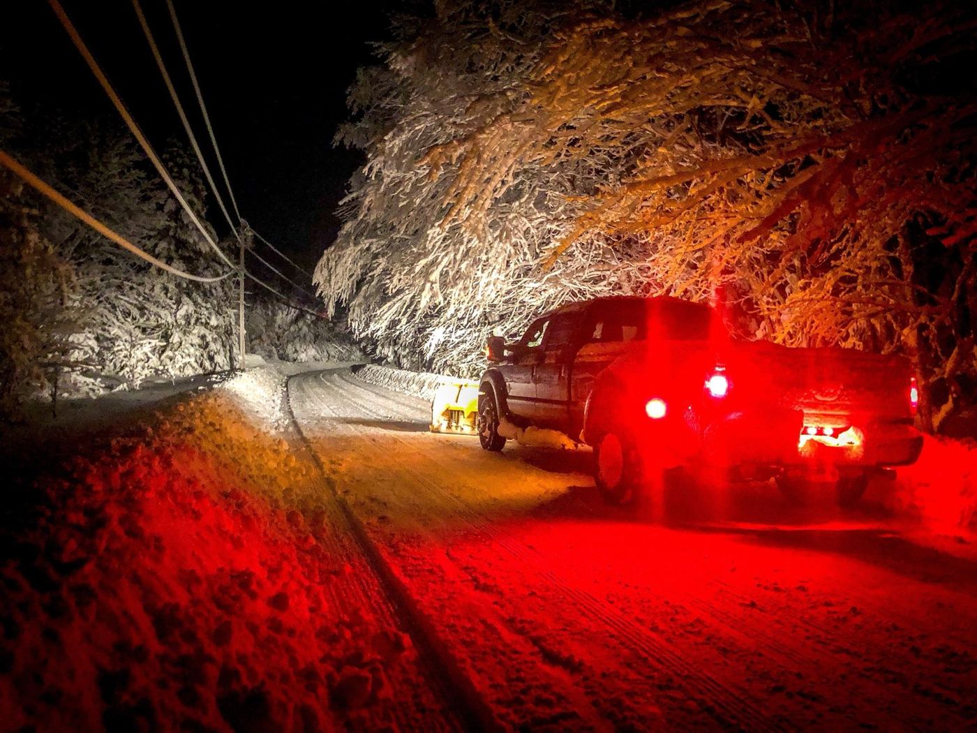 Burgess plow truck clearing a snowy camp road at night under heavy, snow-laden trees
