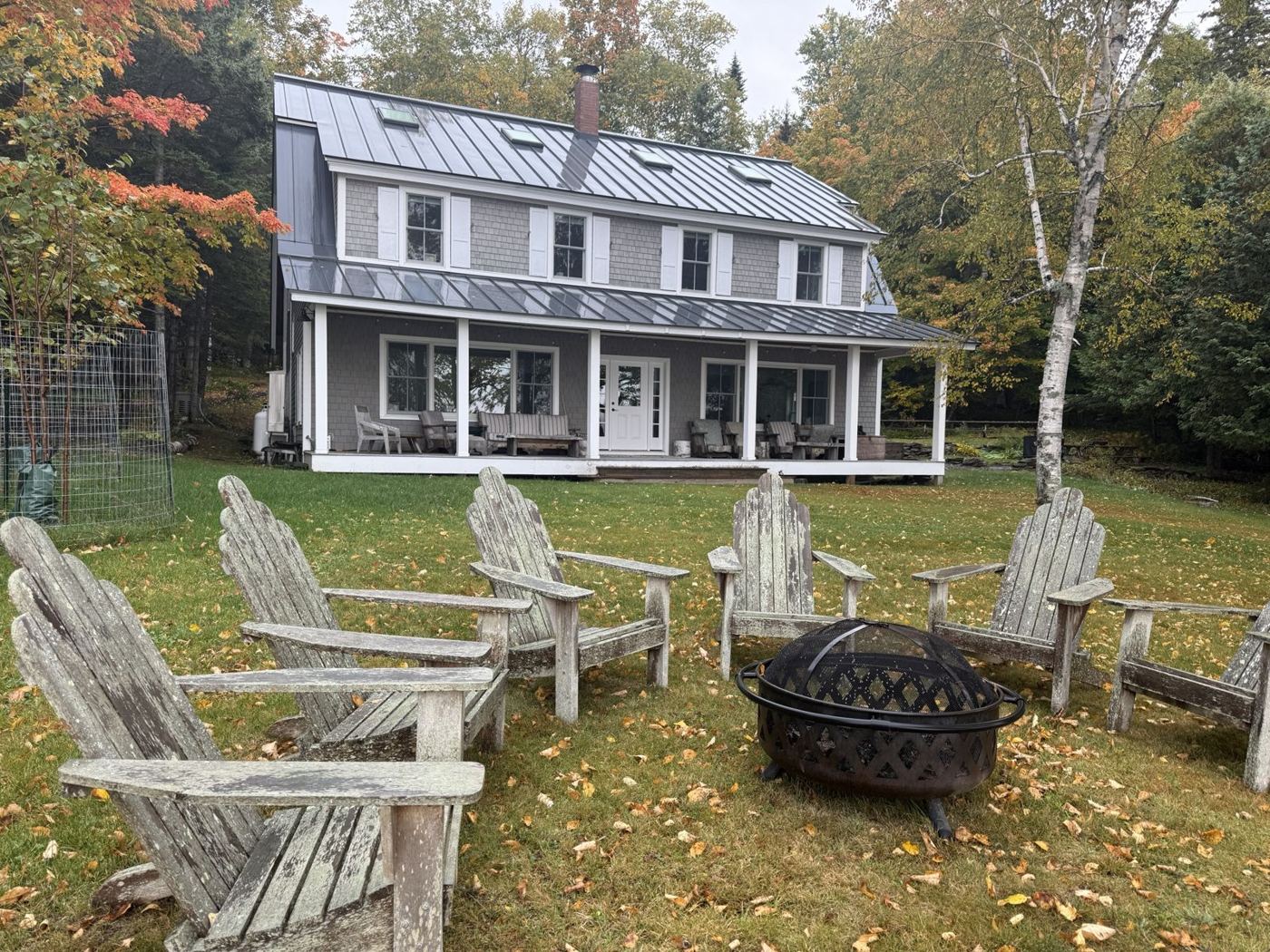 Adirondack chairs around a fire pit on the lawn of a shingled lake house in fall