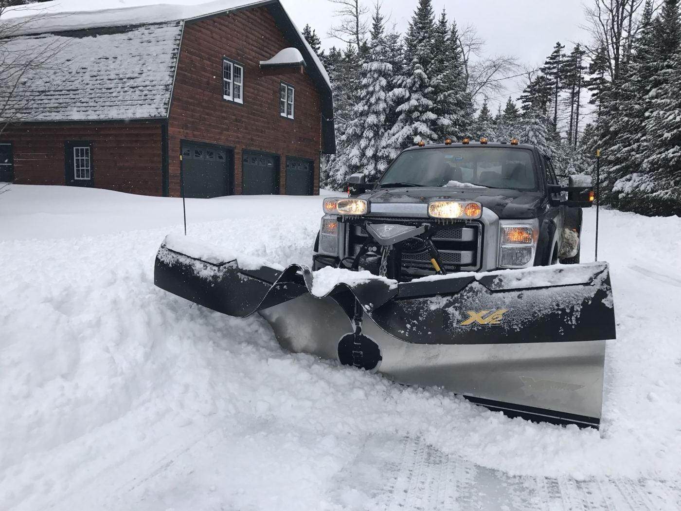 Plow truck with a V-plow clearing the drive in front of a three-bay log garage