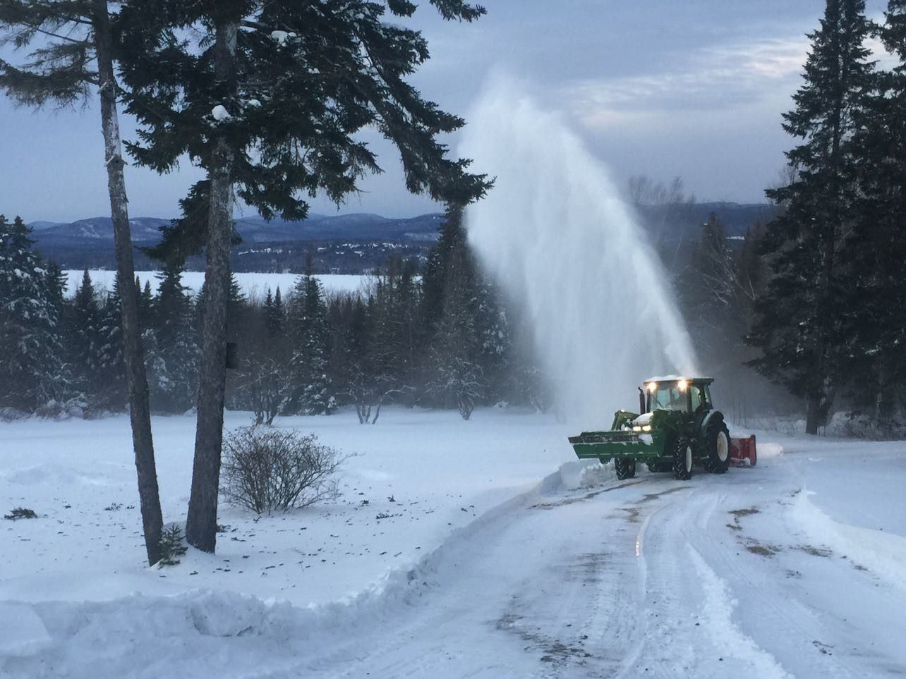 Tractor snowblower clearing a long driveway with Rangeley Lake and the mountains behind